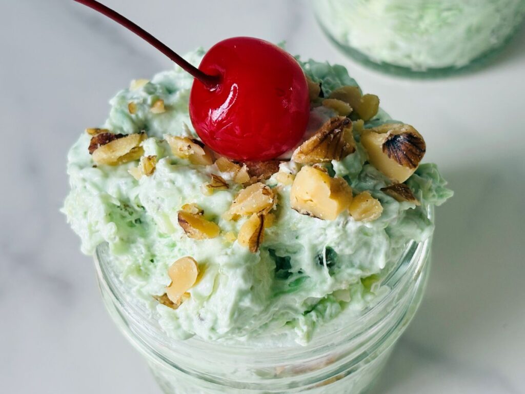 A glass jar filled with a light green creamy dessert, topped with chopped nuts and a bright red maraschino cherry. The jar is set on a white surface.