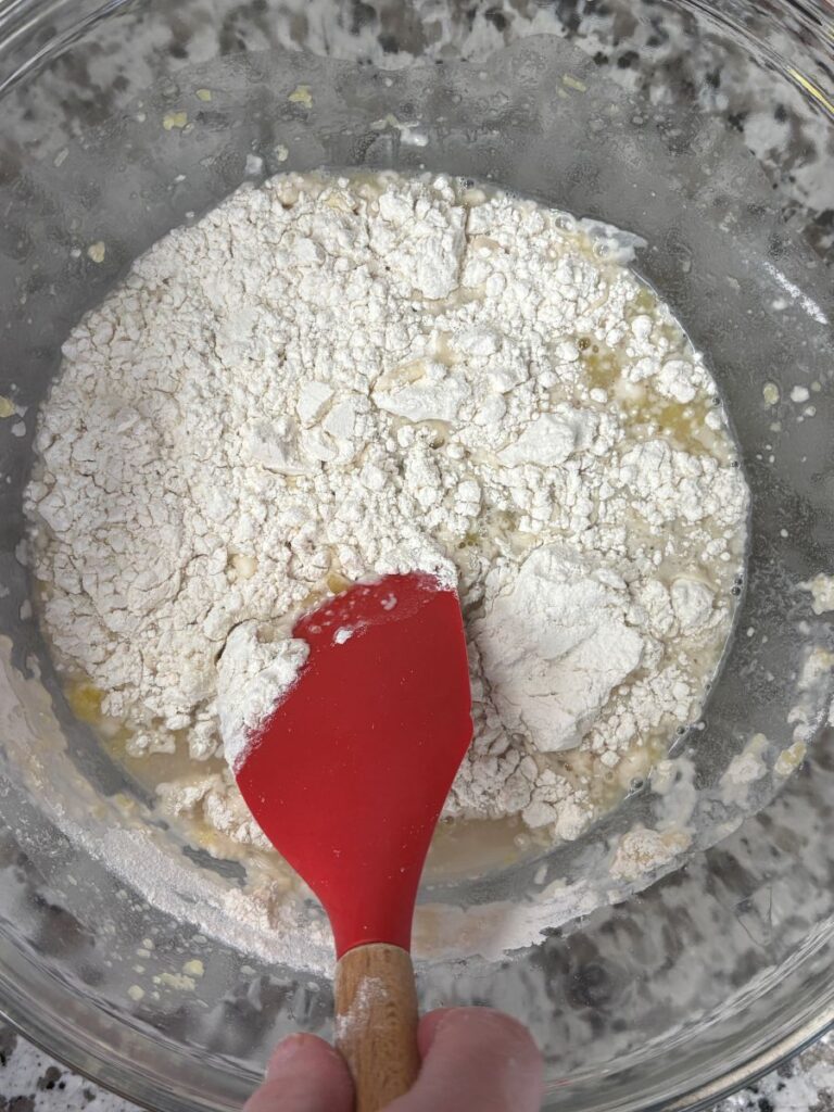 A hand holds a red spatula mixing flour and wet ingredients in a clear glass bowl, preparing dough or batter on a speckled countertop.