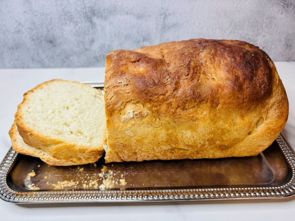 A loaf of bread with one slice cut off, placed on a silver tray against a light gray background.