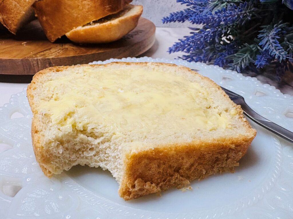 A slice of buttered bread with a bite taken out of it sits on a white decorative plate, next to a knife; more bread and purple flowers are visible in the background.