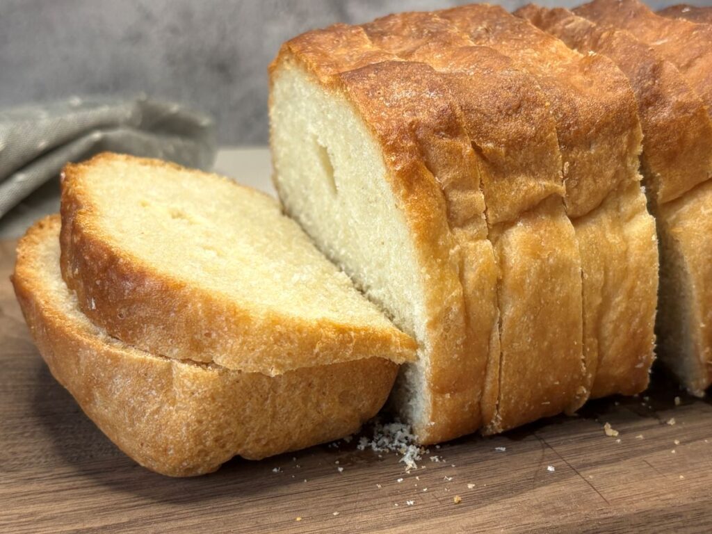 A loaf of golden-brown bread sits on a wooden cutting board, with several slices cut and one slice resting against the loaf. Crumbs are scattered around on the board.