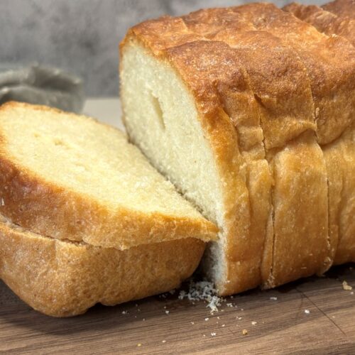 A loaf of golden-brown bread sits on a wooden cutting board, with several slices cut and one slice resting against the loaf. Crumbs are scattered around on the board.