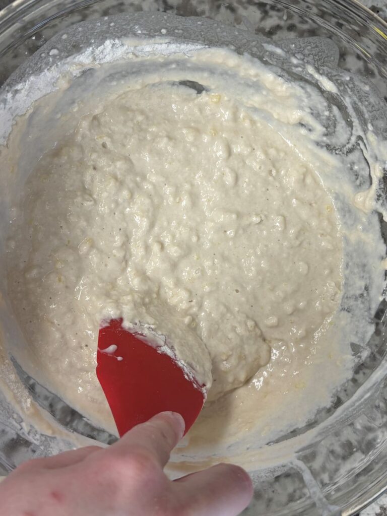 A hand holding a red spatula is mixing a thick, lumpy batter in a glass bowl.