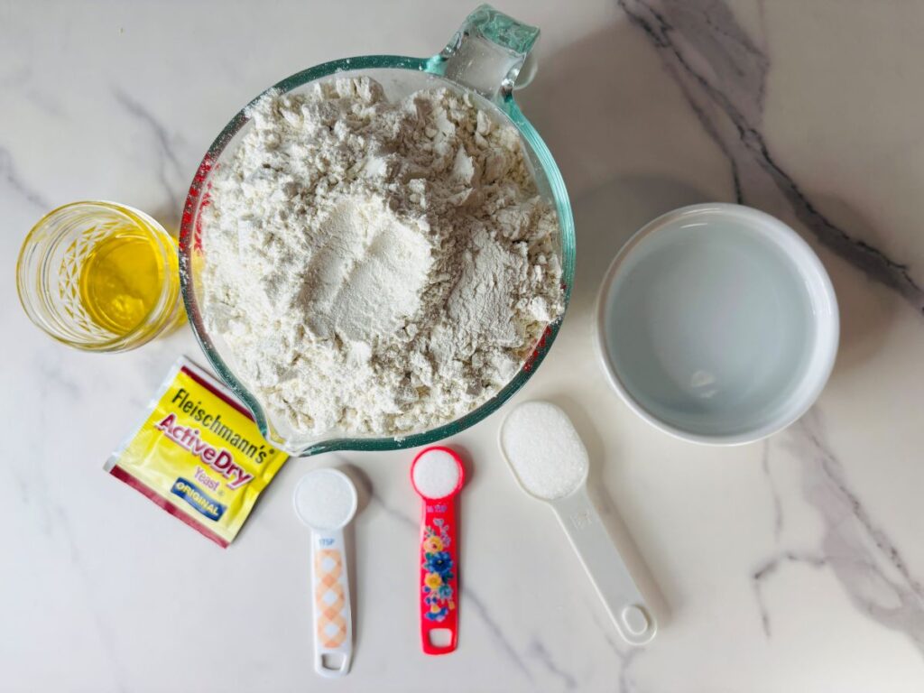 A glass measuring cup of flour, a small bowl of water, a glass with oil, a yeast packet, a tablespoon of sugar, a teaspoon of salt, and a tablespoon of white powder on a marble countertop.