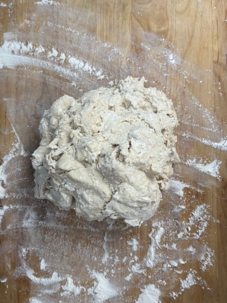 A rough ball of dough sits on a floured wooden surface, surrounded by scattered flour.