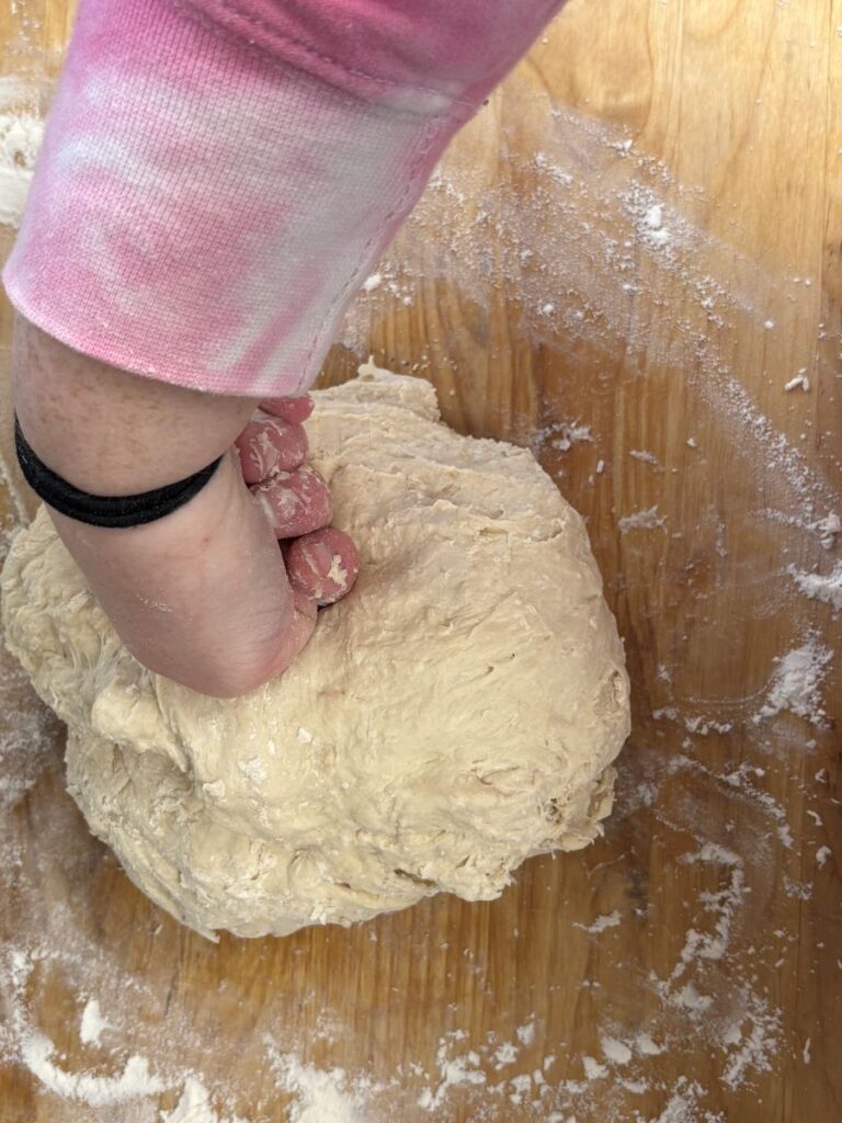 A person wearing a pink and white sleeve kneads a ball of dough on a floured wooden surface.