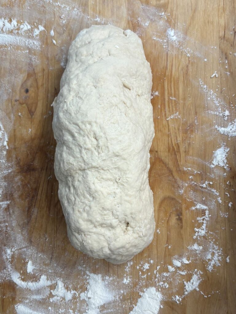 A rough, oblong ball of bread dough rests on a floured wooden surface, with patches of flour scattered around.