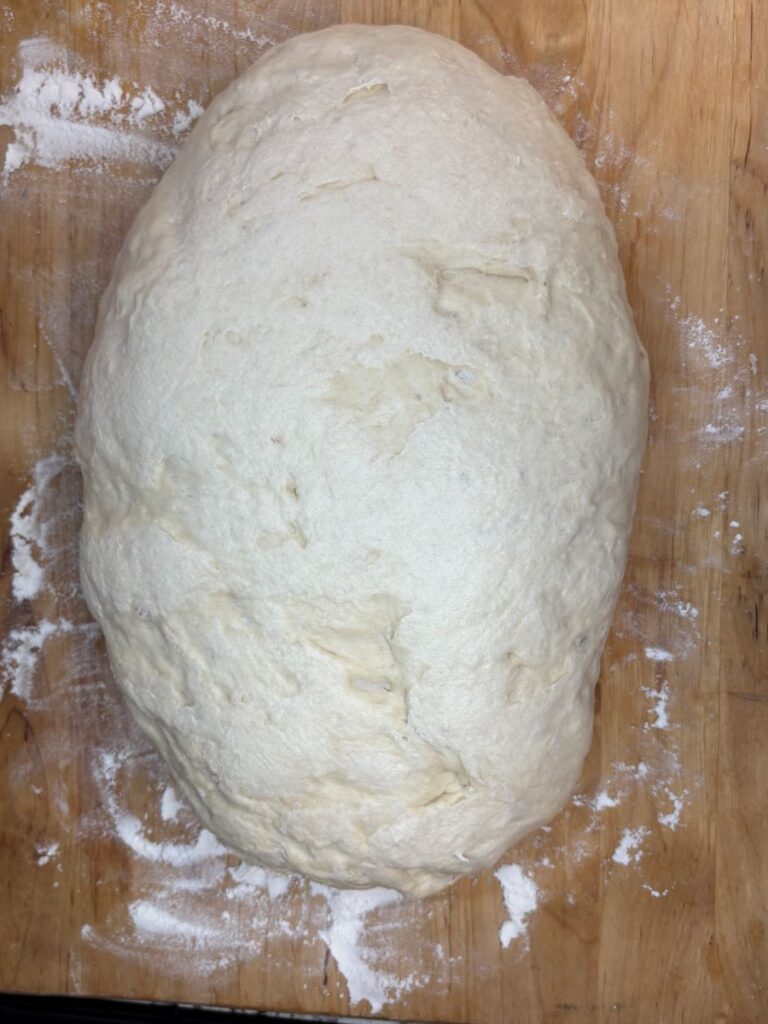 An oval-shaped ball of bread dough rests on a lightly floured wooden surface, ready to be baked.