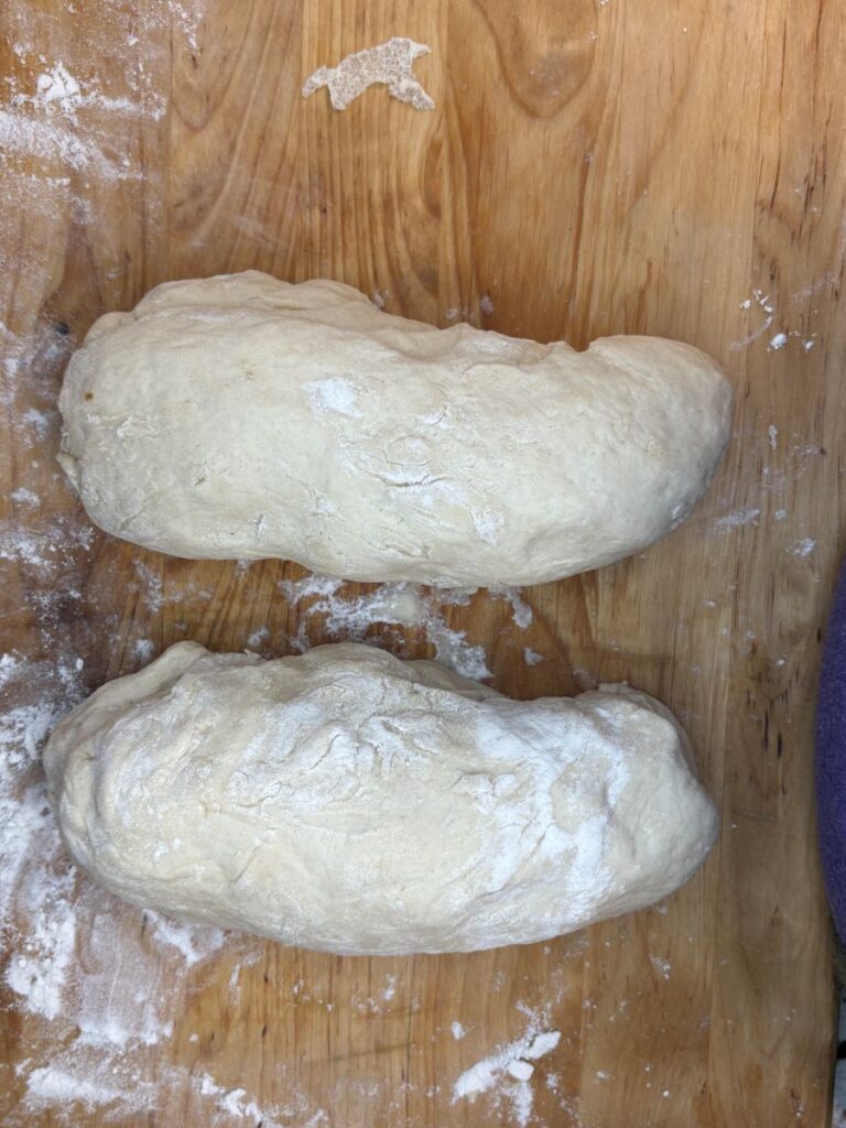 Two oval-shaped pieces of raw bread dough are resting on a floured wooden surface, ready for baking. Flour is scattered around the dough.