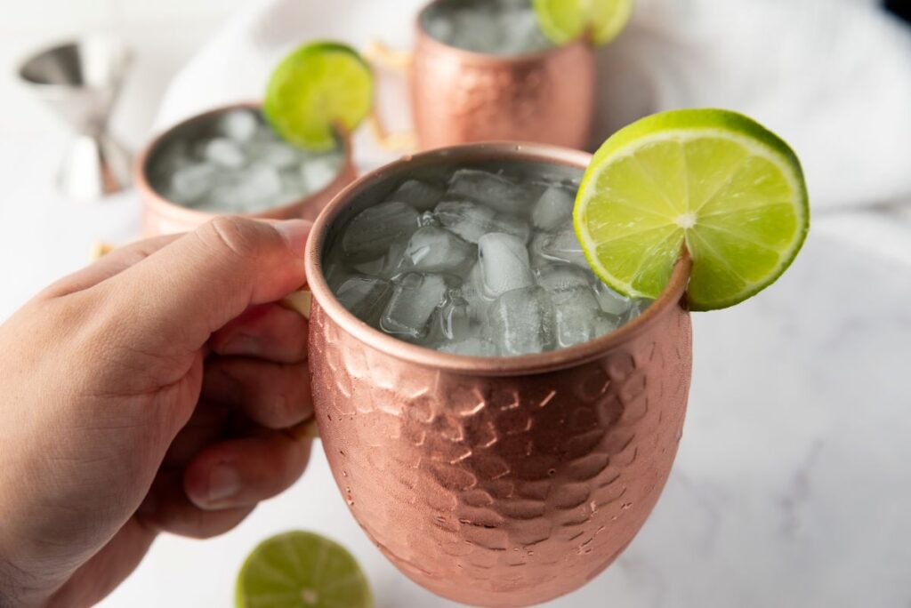 A hand holds a copper mug filled with ice and garnished with a lime slice, with two similar mugs and more lime slices in the background on a white surface.