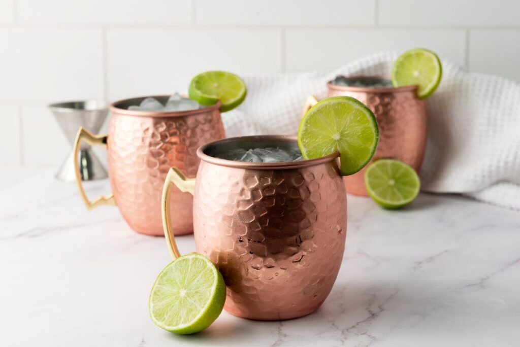 Three copper mugs filled with ice, each garnished with a lime wheel, are set on a white marble surface with a white cloth and a sliced lime nearby. The background features white tile.