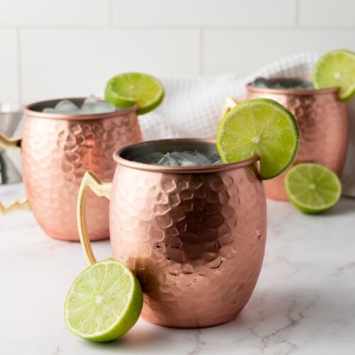 Three copper mugs filled with ice, each garnished with a lime wheel, are set on a white marble surface with a white cloth and a sliced lime nearby. The background features white tile.