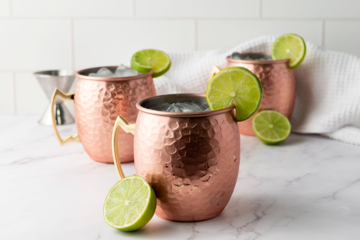 Three copper mugs filled with ice, each garnished with a lime wheel, are set on a white marble surface with a white cloth and a sliced lime nearby. The background features white tile.