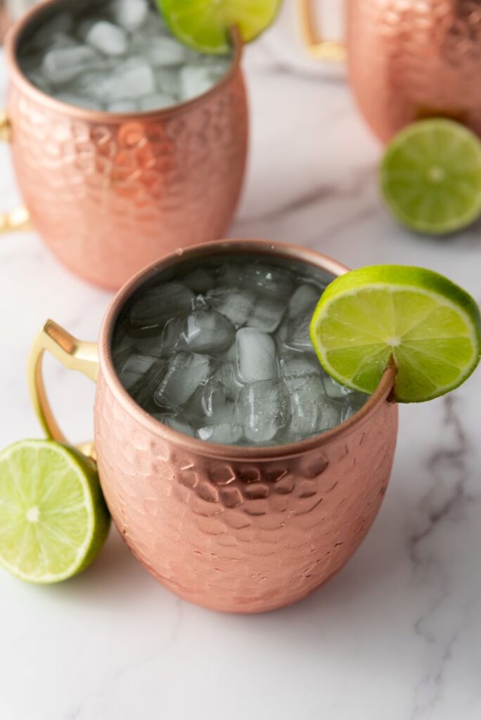 A copper mug filled with ice and a clear drink, garnished with a slice of lime on the rim. Other copper mugs and halved limes are visible in the background on a white marble surface.