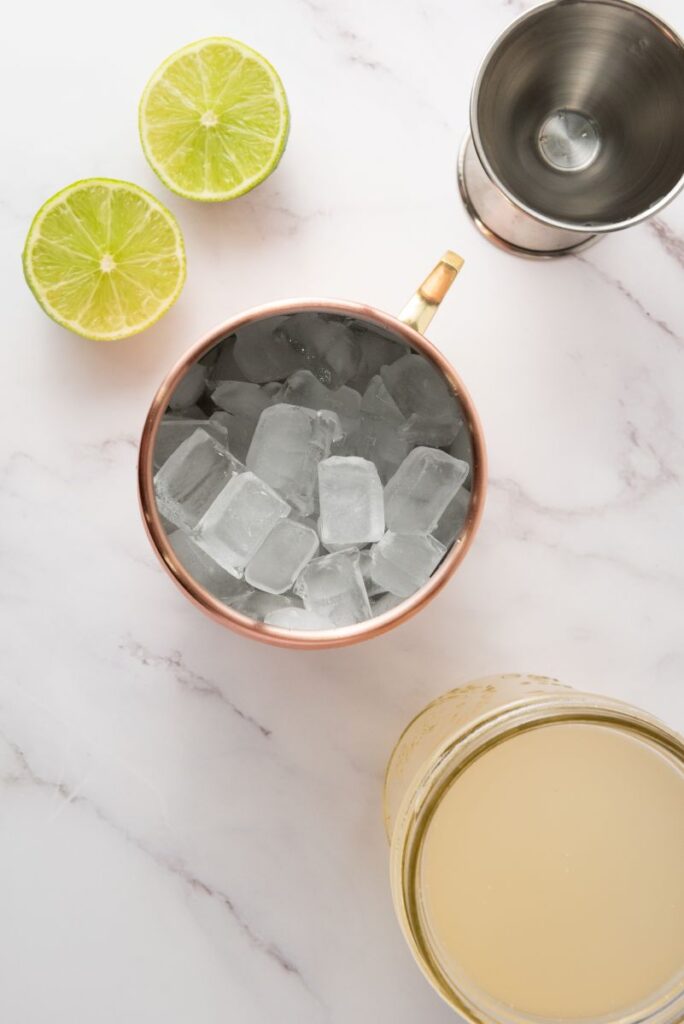Copper mug filled with ice cubes, two lime halves, a jigger, and a jar of pale liquid on a white marble surface.