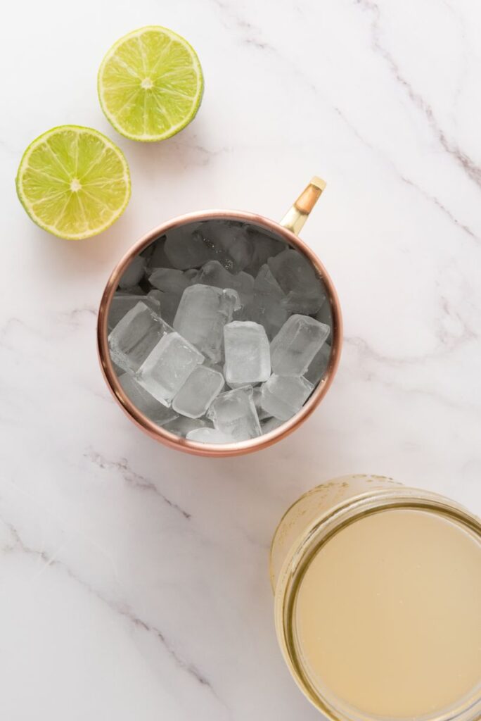 A copper cup filled with ice cubes sits on a marble surface next to a halved lime and a glass jar containing a light yellow liquid.
