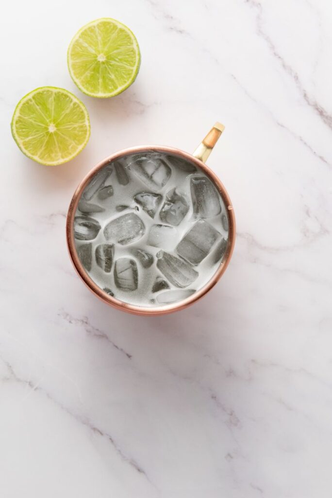 A copper mug filled with a drink and ice cubes sits on a marble surface, next to two halves of a lime.
