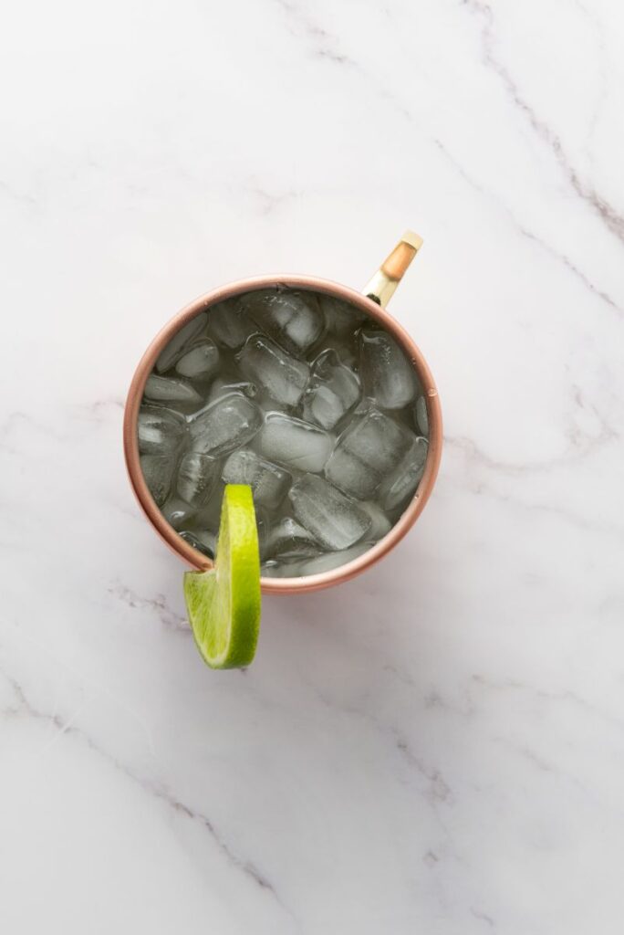 A copper mug filled with ice cubes and a clear drink, garnished with a slice of lime on the rim, sits on a white marble surface.