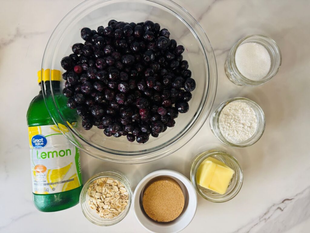 A large bowl of blueberries is surrounded by a bottle of lemon juice, jars of sugar, flour, and oats, a small bowl of brown sugar, and a dish of butter on a marble countertop.