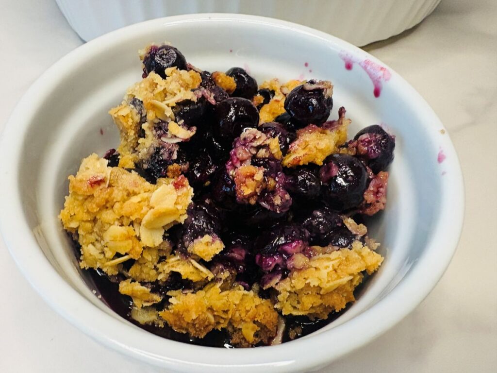 A small white bowl filled with a serving of blueberry crumble, featuring baked blueberries and a golden, crumbly topping.