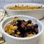 A white bowl filled with blueberry crisp topped with oat crumble sits in front of a larger baking dish of the same dessert. A checkered kitchen towel is beside them on a white countertop.