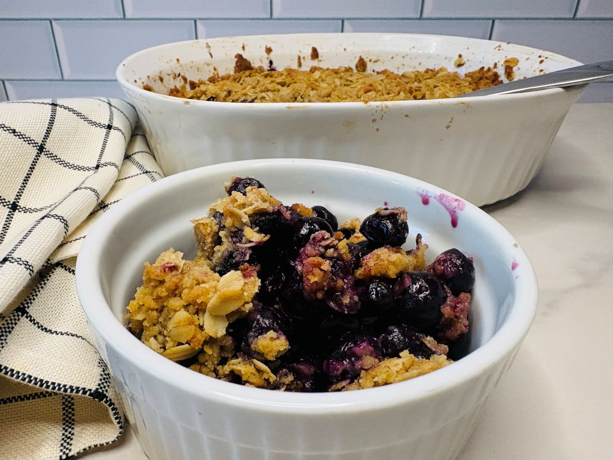 A white bowl filled with blueberry crisp topped with oat crumble sits in front of a larger baking dish of the same dessert. A checkered kitchen towel is beside them on a white countertop.
