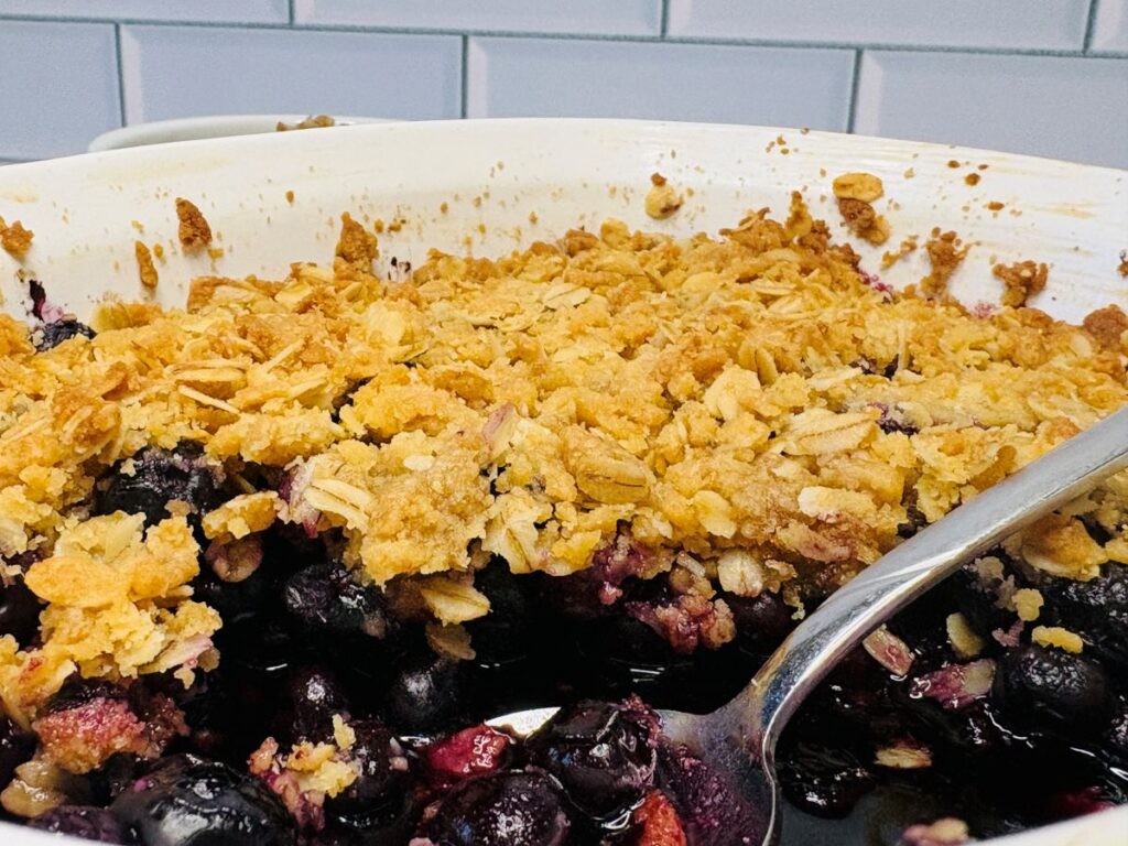 A close-up of a blueberry crisp in a white baking dish, showing a golden oat crumb topping and juicy blueberries underneath. A silver serving spoon is resting inside the dish. White tile backsplash is in the background.