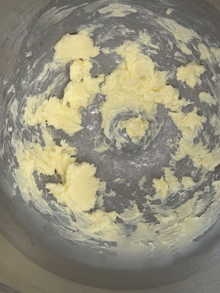 Softened, creamy butter is partially mixed and spread around the sides and bottom of a large stainless steel mixing bowl, showing a whipped, smooth texture.