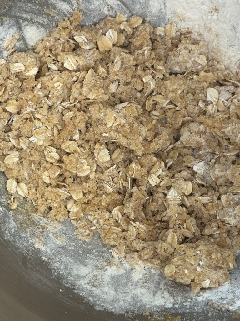 A close-up of an oat and flour mixture in a mixing bowl, showing rough, crumbly texture with visible rolled oats and patches of dry flour.