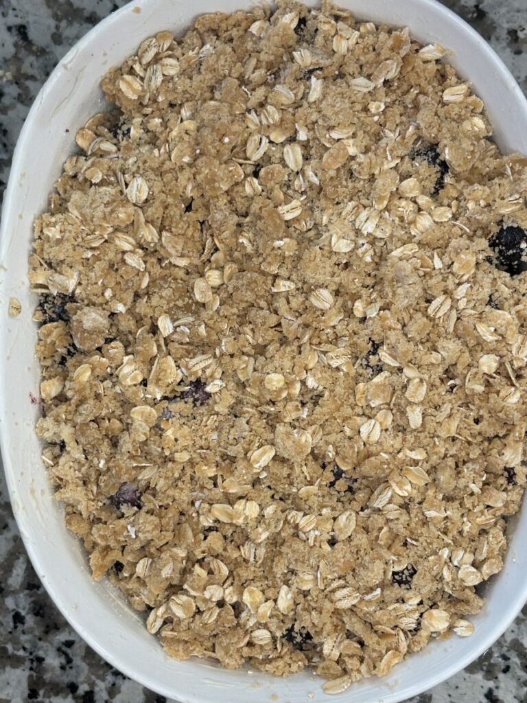 A close-up of an unbaked fruit crisp in a white oval dish, topped with a crumbly oat and brown sugar mixture, set on a speckled granite countertop.
