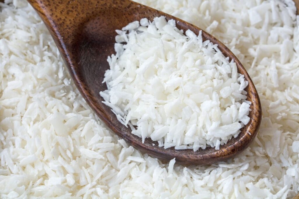 A close-up of a wooden spoon filled with shredded coconut, resting on a surface covered with more shredded coconut.