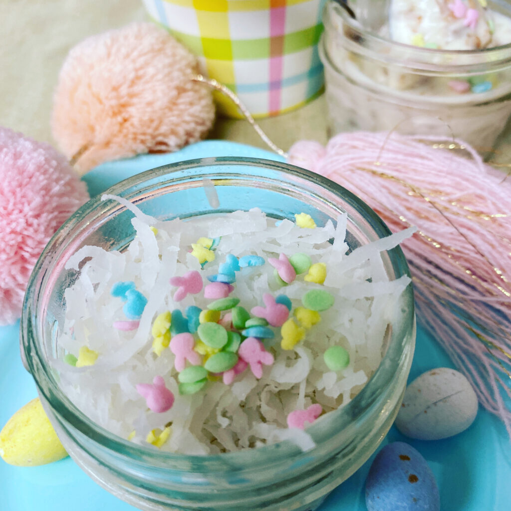 A small glass jar filled with shredded coconut and colorful pastel sprinkles sits on a blue plate, surrounded by Easter eggs, pink yarn pom-poms, and another dessert jar in the background.