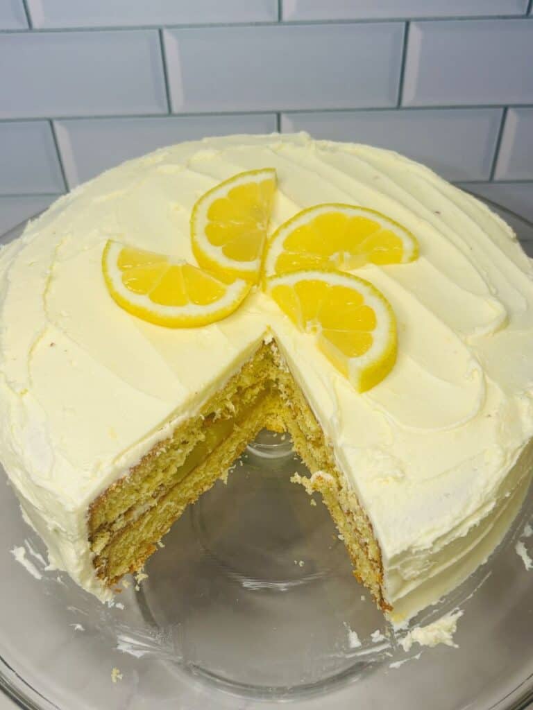 A round lemon layer cake with creamy white frosting, topped with three lemon slices, sits on a glass cake stand. A large slice is missing, revealing the moist layers inside. The background is white subway tile.