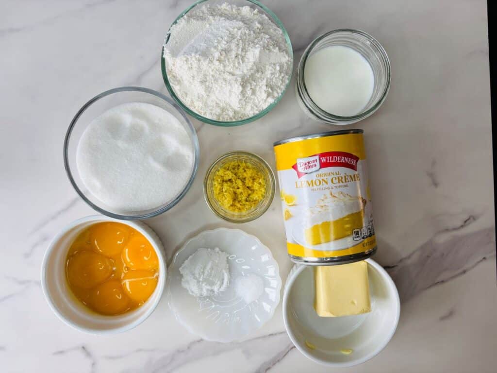 Baking ingredients on a white surface: a bowl of flour, a bowl of sugar, a jar of milk, a jar of lemon zest, a bowl of egg yolks, a can of lemon crème, a dish of butter, and a plate with baking powder and baking soda.