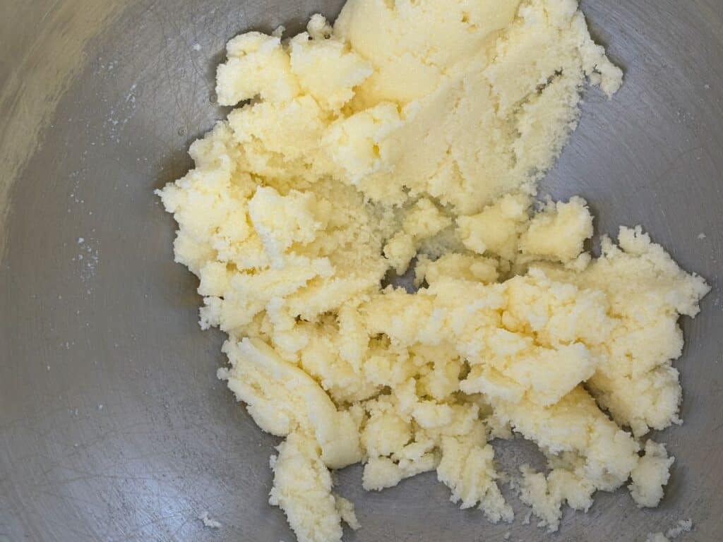 A close-up view of creamed butter and sugar in a metal mixing bowl, showing a light, fluffy texture with pale yellow color.