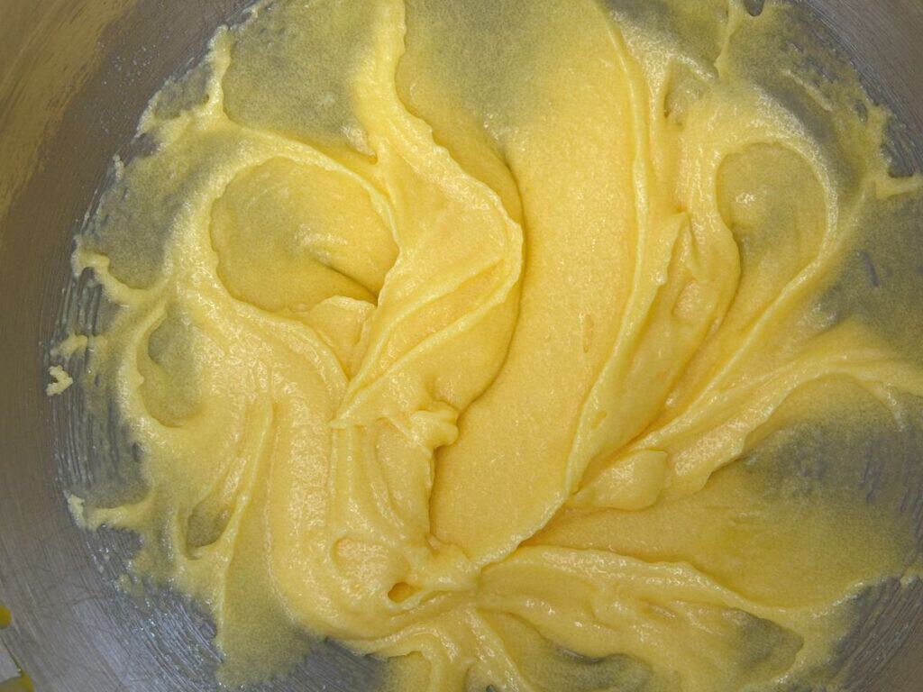 Smooth, creamy yellow batter is spread in swirling patterns inside a metal mixing bowl, suggesting it is in the process of being mixed for baking.