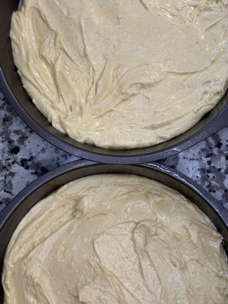 Two round cake pans filled with raw, smooth yellow cake batter, sitting on a granite countertop, ready to be baked.