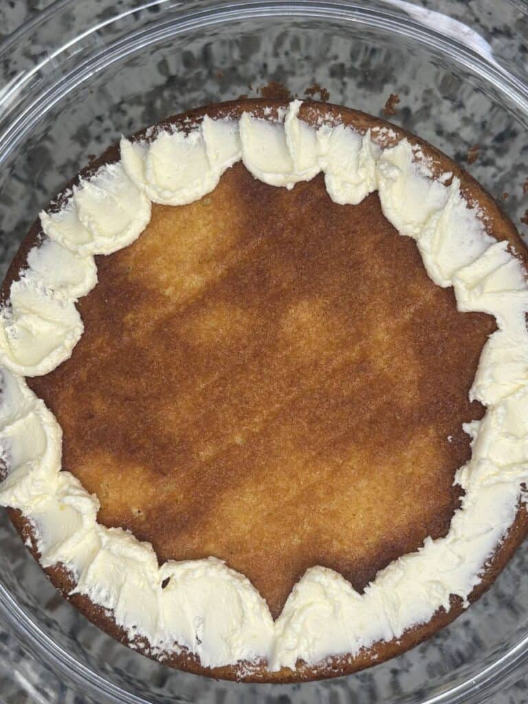 A round, plain sponge cake sits on a glass plate, decorated with a simple ring of white buttercream icing around the edge. The cake has a golden brown top and no additional toppings or decorations.