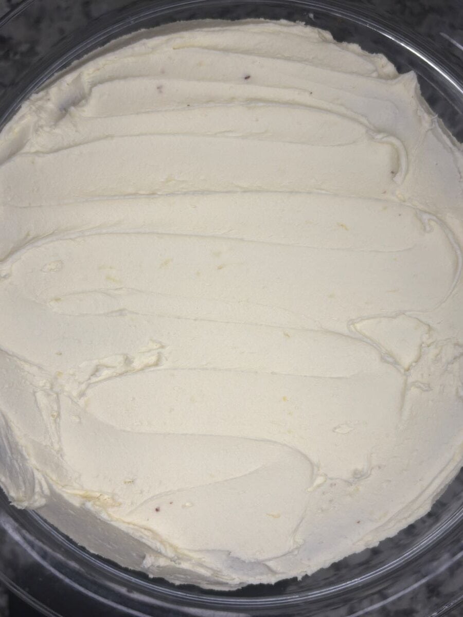 A close-up of a container filled with smooth, creamy white frosting or whipped cream, evenly spread with visible swirled lines on the surface.