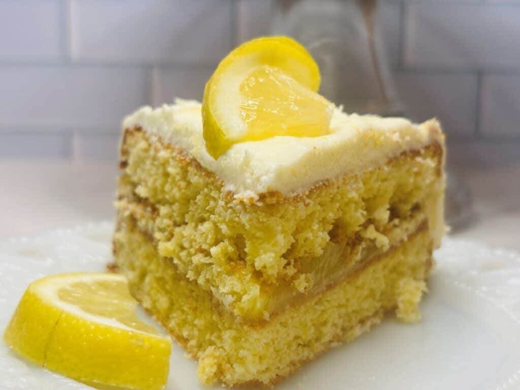 A slice of layered lemon cake with creamy frosting is topped with a lemon wedge, sitting on a white plate. Another lemon wedge rests beside the cake. The background features a white tiled wall.