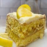 A slice of layered lemon cake with creamy frosting is topped with a lemon wedge, sitting on a white plate. Another lemon wedge rests beside the cake. The background features a white tiled wall.
