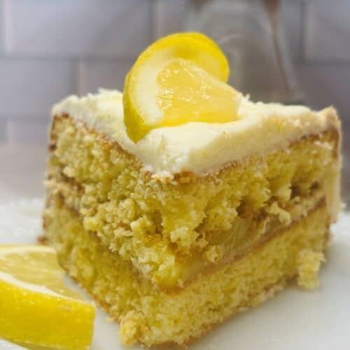 A slice of layered lemon cake with creamy frosting is topped with a lemon wedge, sitting on a white plate. Another lemon wedge rests beside the cake. The background features a white tiled wall.