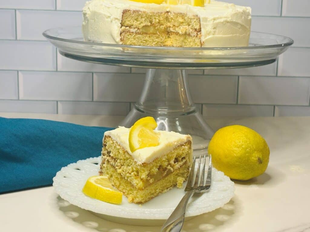 A slice of lemon cake with white frosting and a lemon wedge sits on a white plate with a fork. Behind it, the rest of the cake is displayed on a glass stand. A whole lemon and a blue napkin are nearby.