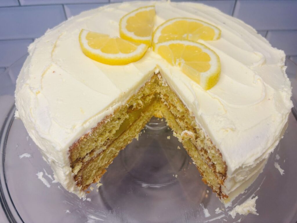 A three-layer lemon cake with white frosting, topped with lemon slices, sits on a glass cake stand. A large wedge has been cut out, revealing the cakes moist, yellow interior and creamy layers.