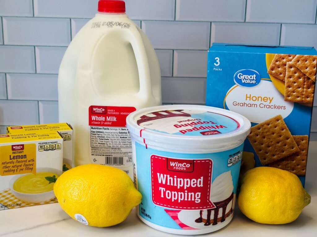 A container of whole milk, a box of honey graham crackers, a tub of whipped topping, a box of lemon pudding mix, and two fresh lemons are arranged on a kitchen counter with a tiled backsplash.