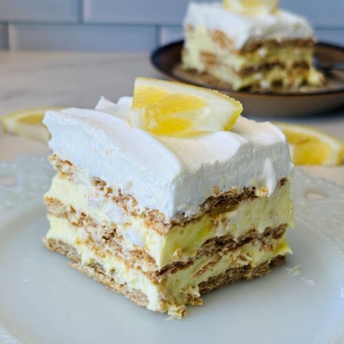 A slice of lemon icebox cake with layers of whipped cream, graham crackers, and lemon filling, topped with a lemon wedge, sits on a white plate with lemon slices in the background.