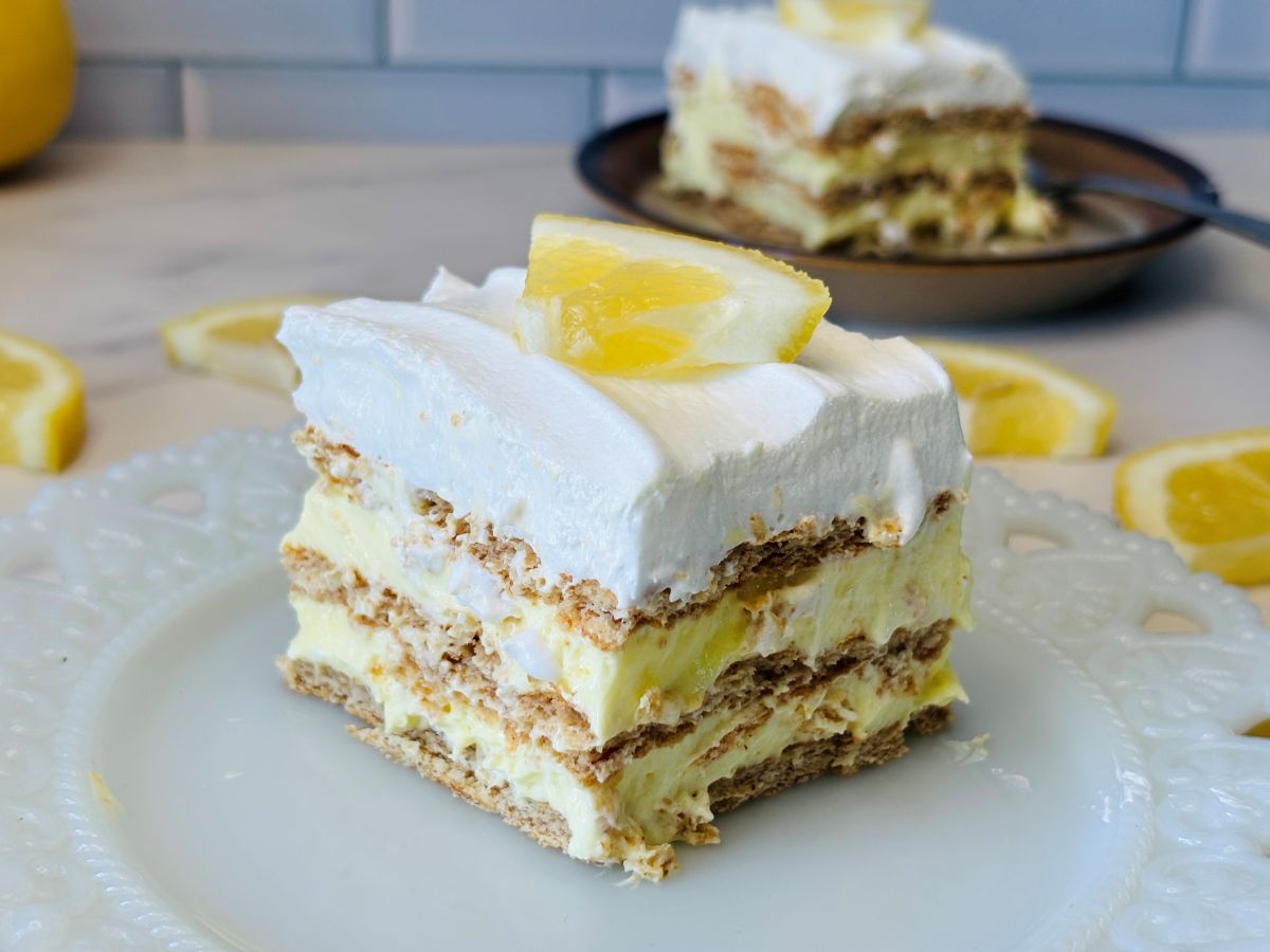 A slice of lemon icebox cake with layers of whipped cream, graham crackers, and lemon filling, topped with a lemon wedge, sits on a white plate with lemon slices in the background.