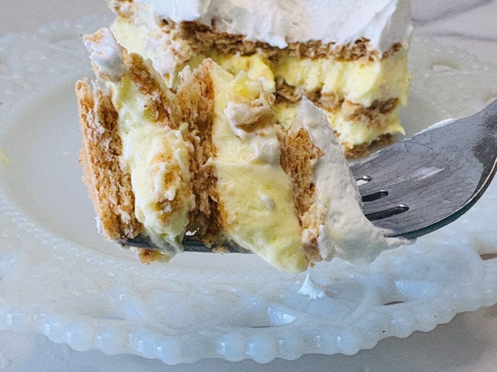 A close-up of a fork holding a bite of layered dessert with graham crackers, creamy yellow custard, and whipped topping, with the rest of the dessert on a white ornate plate in the background.