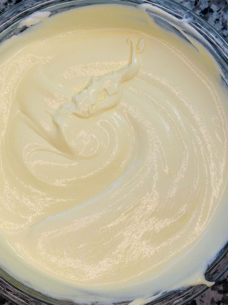 A close-up of a bowl filled with smooth, creamy, pale yellow batter or frosting, swirled in a circular pattern. The surface has a glossy texture and is set on a speckled countertop.