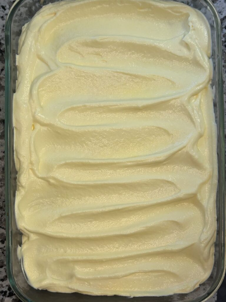 A rectangular glass dish filled with a dessert topped with thick, smooth, creamy white frosting, swirled into gentle waves. The dish is placed on a gray speckled countertop.